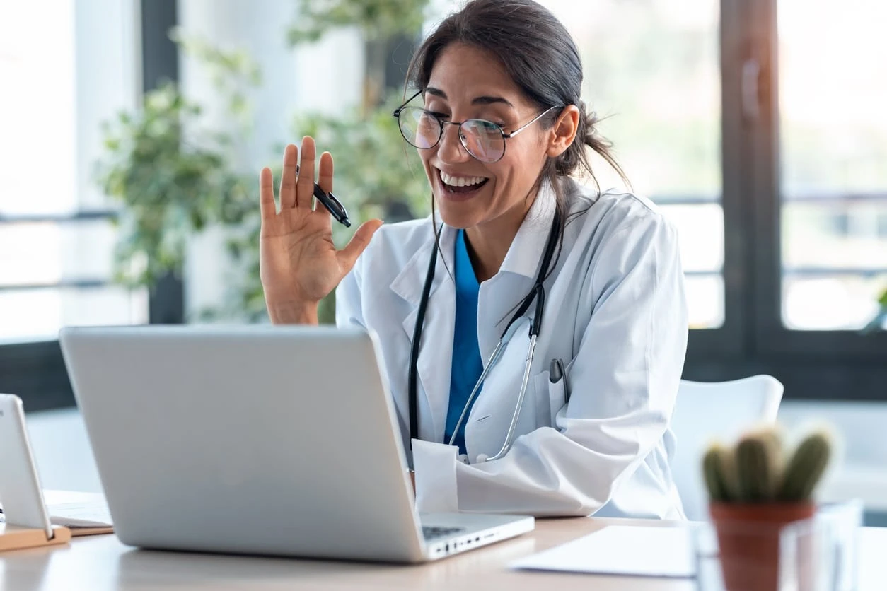 Seorang dokter wanita mengenakan jas putih dan kacamata, tersenyum sambil melambaikan tangan di depan laptop.